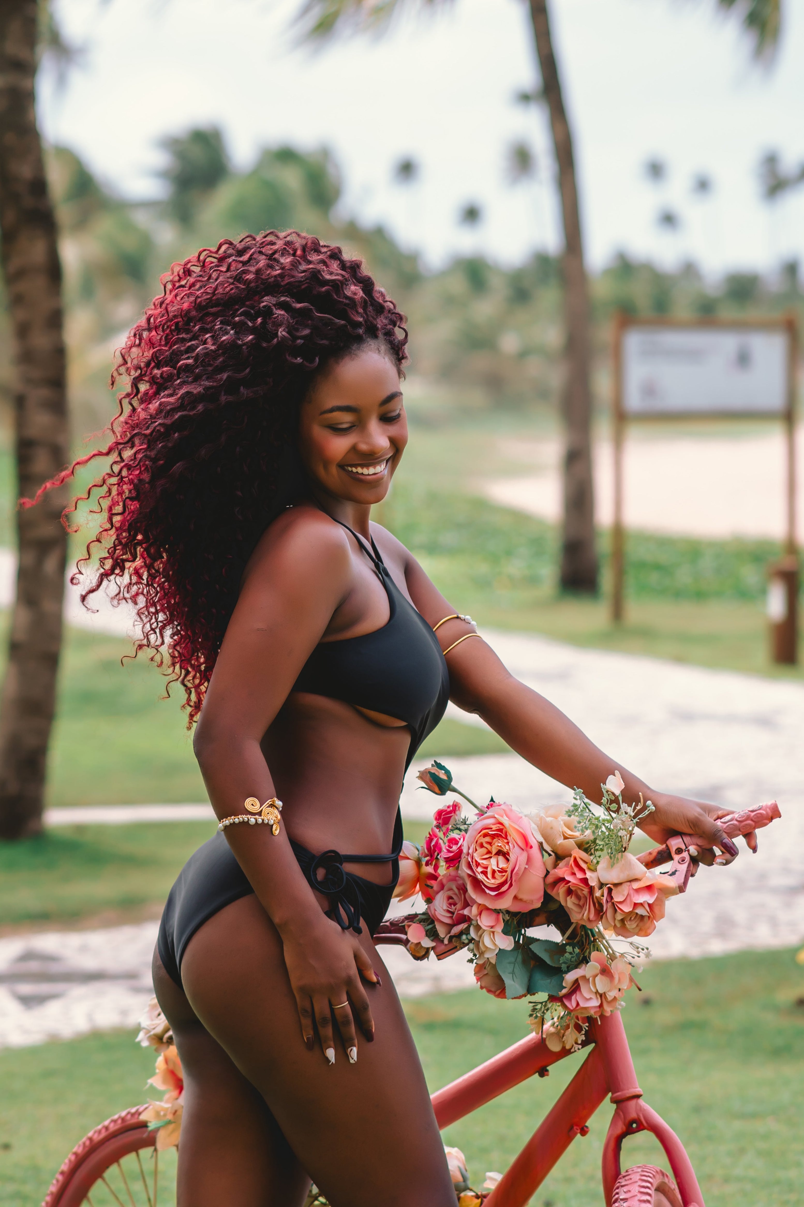 Woman with curly hair, wearing The Sway asymmetrical, dig-free one-piece swimsuit in Black, standing next to a bicycle with flowers in a tropical setting